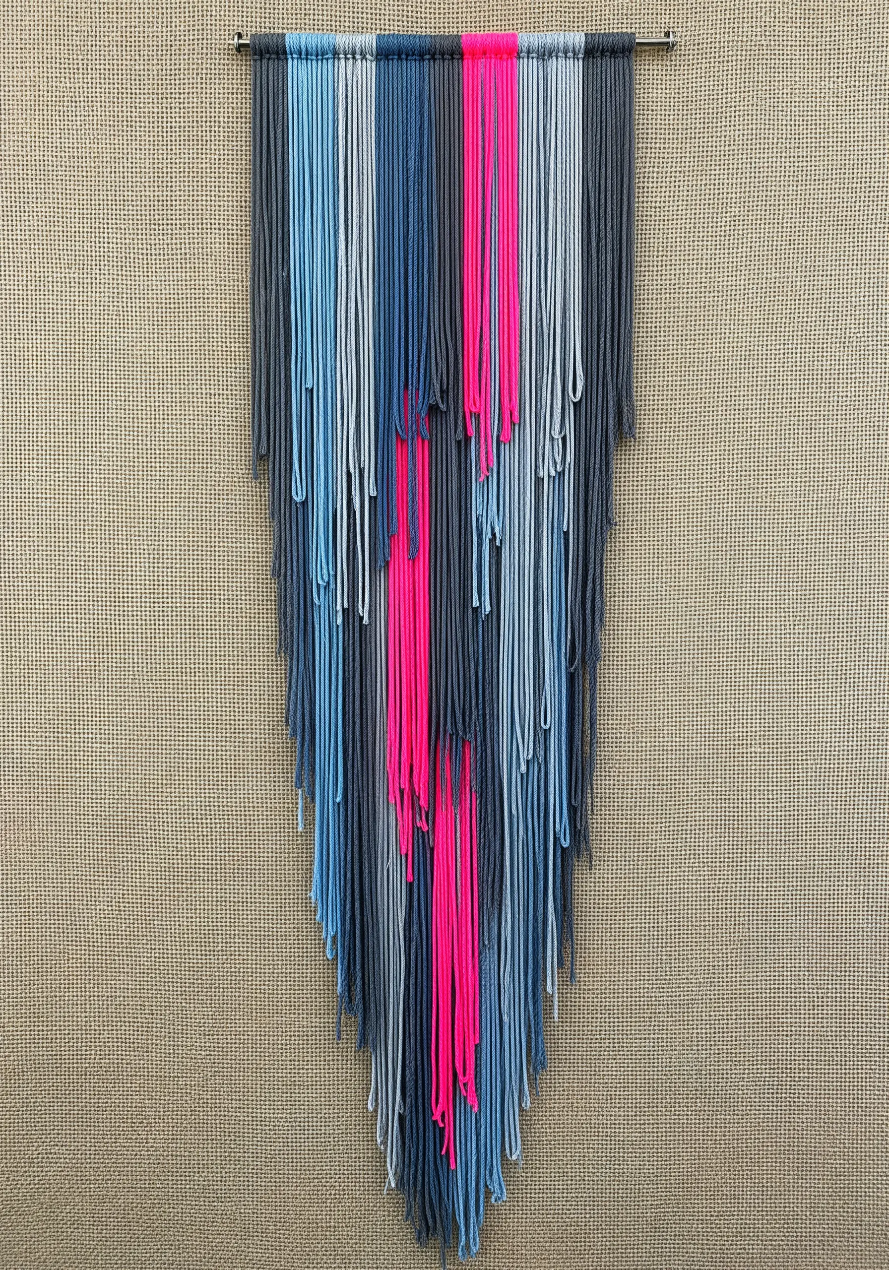 A fiber wall hanging with various shades of blue and gray, featuring a single, vibrant streak of neon pink.