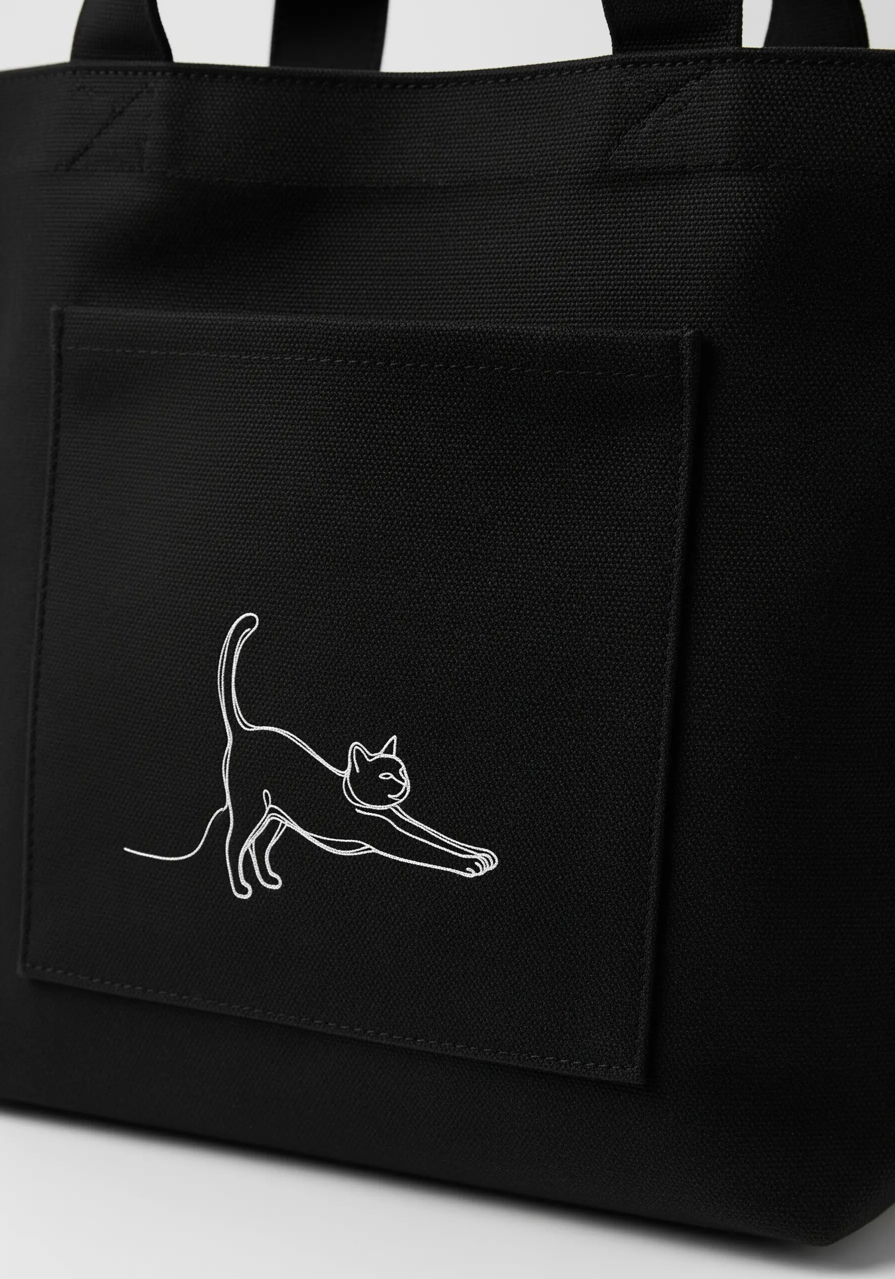 A single-line drawing of a stretching cat embroidered in white on the pocket of a black tote bag.