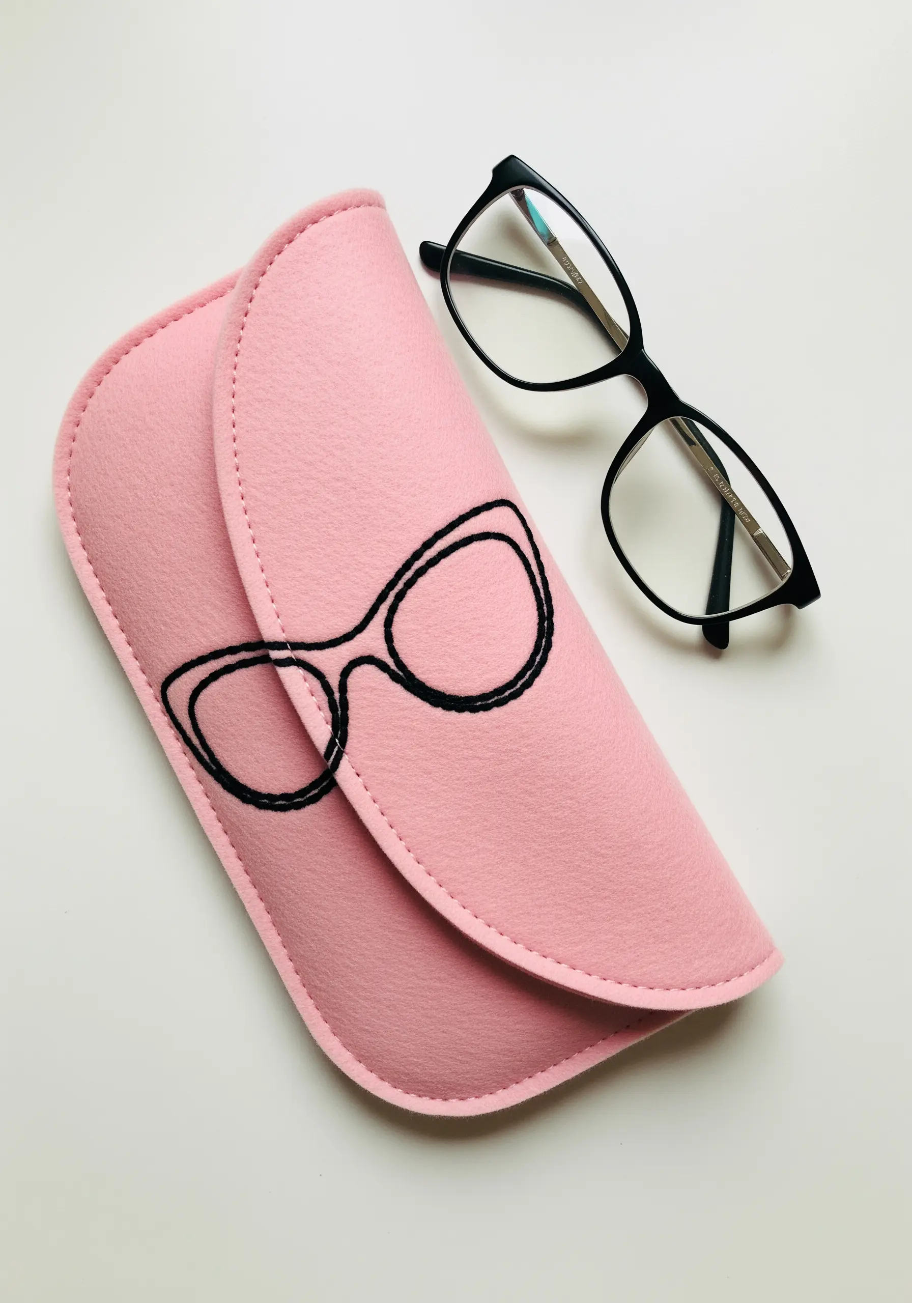 A pink felt glasses case embroidered with the simple black outline of a pair of cat-eye glasses.